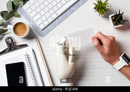 Immagine ritagliata di imprenditore con mano cyborg e smartwatch holding blank carta da visita a tavola con lo smartphone con schermo vuoto in ufficio Foto Stock