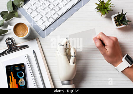 Immagine ritagliata di imprenditore con mano cyborg e smartwatch holding blank carta da visita a tavola con lo smartphone con una infografica sullo schermo in office Foto Stock