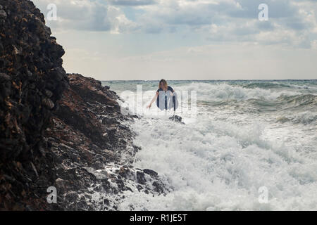 Turista arrampicata sulle rocce al mare tempestoso con grandi onde, schizzi e schiuma. Disperato e pericoloso atto. Foto Stock