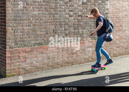 Donna anziana lo skateboard passato un muro di mattoni su una rosa e verde del bambino skateboard, London, Regno Unito Foto Stock