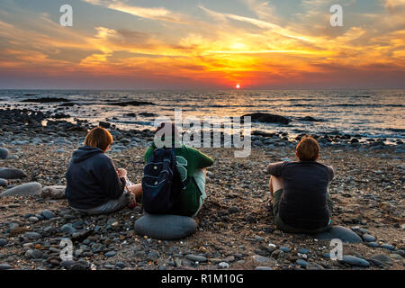 Famiglia di tre persone (madre, figlio, figlia) che si siede su una spiaggia di pietra guardando il tramonto a Westward ho!, Devon, Regno Unito Foto Stock