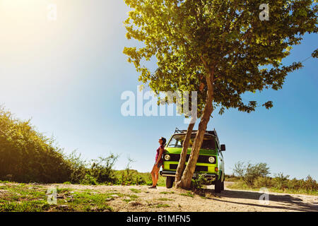 Giovane e bella ragazza in pantaloncini corti in piedi accanto al vecchio classico timer camper van sotto l'albero su un luminoso e caldo giorno d'estate Foto Stock