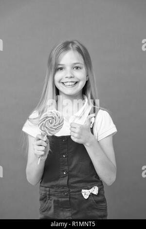 Ragazza di mangiare lecca-lecca colorati. Infanzia dolce concetto. Il capretto con capelli lunghi ama i dolci. Ragazza con volto sorridente detiene caramella gigante in mano, sfondo violetto. Bambino in tuta rosa che mostra il pollice verso l'alto. Foto Stock