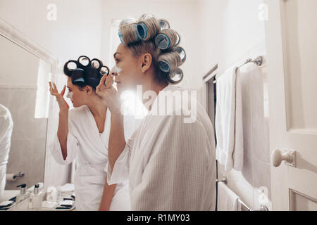 Vista laterale di due donne in accappatoi di applicare la crema sulla faccia in piedi di fronte a specchio. Le donne con rulli di ricci appuntato sui capelli toelettatura a casa. Foto Stock