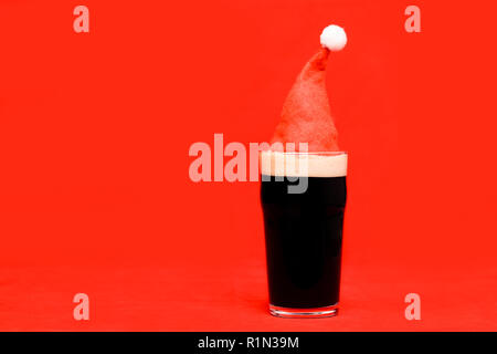 Nonik pinta bicchiere di birra scura o stout ale con red santa hat sul rosso sfondo di Natale Foto Stock