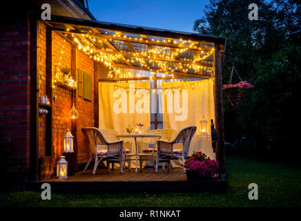 Vista su accogliente terrazza esterna con tavolo e sedie, molto romantica illuminazione, bianco lanterne e candele accese, stringa di led luci di partito e le lampadine con Foto Stock