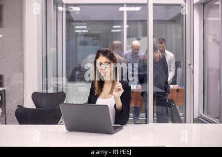 Una giovane donna lavora sul computer portatile in ufficio, gruppo dei lavoratori tavolo da gioco calcio in altro ufficio. Foto Stock