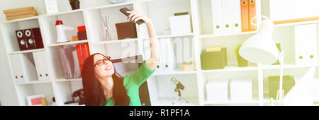 Una giovane ragazza con gli occhiali si siede in ufficio a tavola e scatta foto di se stessa sul telefono. Foto Stock