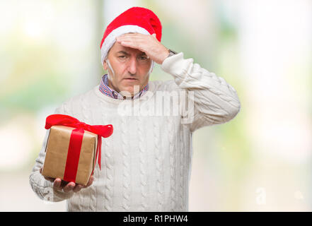 Bello il senior uomo con cappello a Natale e dono di contenimento su sfondo isolato ha sottolineato con la mano sulla testa, sconvolto con vergogna e la sorpresa di fronte, Foto Stock