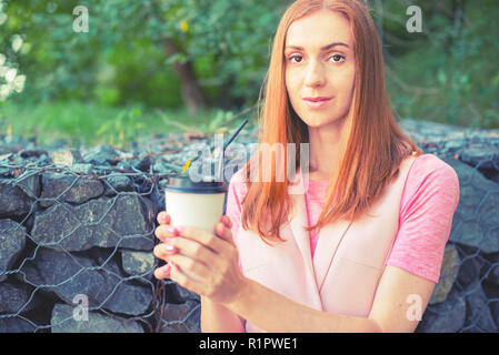 Una bella i Capelli rossi ragazza va giù per le scale e beve caffè da un bicchiere di carta. La prima colazione di stile di vita nel parco Foto Stock