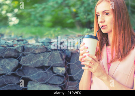 Una bella i Capelli rossi ragazza va giù per le scale e beve caffè da un bicchiere di carta. La prima colazione di stile di vita nel parco Foto Stock