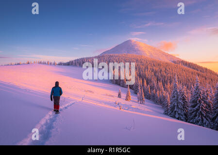 Escursioni d inverno e passeggiate in montagna. Il ragazzo va giù il percorso nella neve. Bel tramonto. Paesaggio con una cima di montagna e foreste di abete rosso Foto Stock