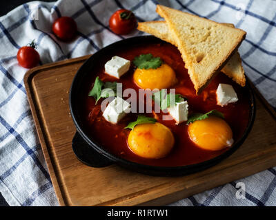 Uova fritte con polpa di pomodoro e formaggio feta in una ghisa pan su un tagliere di legno Foto Stock
