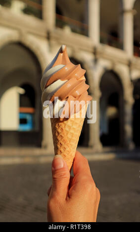 Donna di mano che tiene un due-tono Soft servire il gelato cono, con sfocate edificio Vintage in background Foto Stock