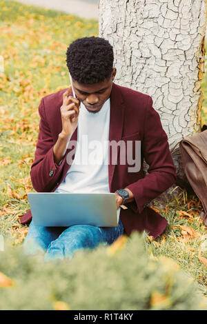 African American telelavoratore utilizzando laptop e parlando sullo smartphone in posizione di parcheggio Foto Stock