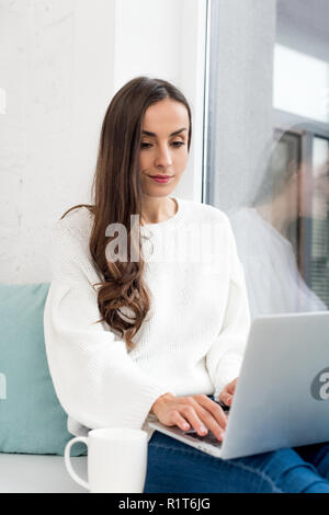 Giovane e bella freelancer con tazza di caffè a lavorare con il computer portatile sul davanzale di casa Foto Stock