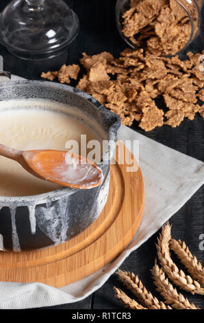 Latte bollito in una pentola rustico, oltre rovesciato sopra i bordi e ribaltato un vasetto con cereali di grano. La colazione tavolo rustico. Mattina di routine. Foto Stock