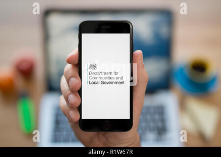 Un uomo guarda al suo iPhone che visualizza il Dipartimento per le comunità e il governo locale logo, mentre se ne sta seduto alla sua scrivania per computer (solo uso editoriale) Foto Stock