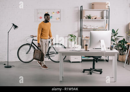 Bello African American designer in arancione maglione entrando in ufficio con valigetta Foto Stock