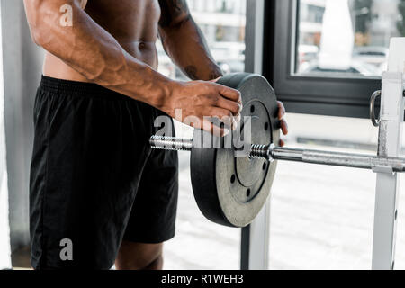 Ritagliato colpo di muscolare sportivo giovani mettendo la piastra di peso su barbell in palestra Foto Stock