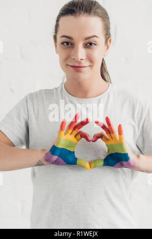 Sorridente giovane uomo transgender facendo segno di cuore con le mani nei colori della bandiera di orgoglio di fronte bianco muro di mattoni Foto Stock