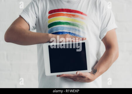 Ritagliato colpo di uomo in t-shirt con orgoglio bandiera tenendo tablet con schermo vuoto di fronte bianco muro di mattoni Foto Stock