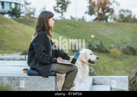 Vista laterale del freelancer strofinarsi il cane e utilizzando laptop in posizione di parcheggio Foto Stock