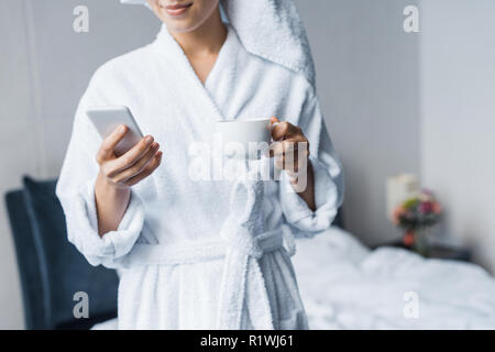Vista ritagliata della donna in accappatoio holding tazza di caffè e utilizza lo smartphone in camera da letto al mattino Foto Stock