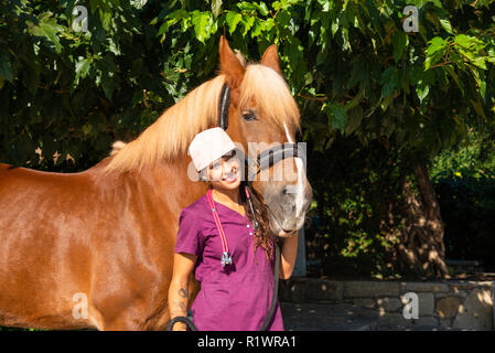 Sorridente giovane femmina medico veterinario ritratto con cavallo marrone all'aperto. Assistenza sanitaria equestre concetto medico. Foto Stock