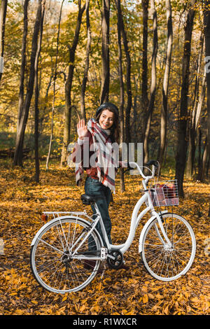 Sorridente ragazza elegante in giacca di pelle e beret agitando a mano e il trasporto bicicletta nel parco autunnali Foto Stock