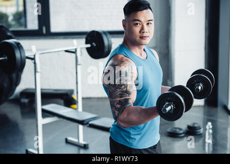 Concentrato di giovani asiatici sportivo esercizio con manubri in palestra e guardando la fotocamera Foto Stock