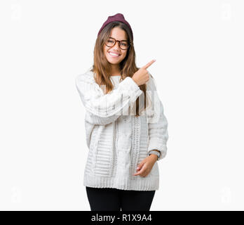 Giovane bella bruna hipster donna che indossa occhiali e cappello invernale su sfondo isolato allegro con un sorriso della faccia rivolta con la mano e fi Foto Stock