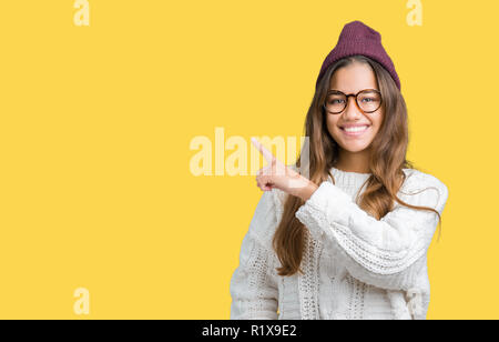 Giovane bella bruna hipster donna che indossa occhiali e cappello invernale su isolati di puntamento dello sfondo con la mano il dito a lato mostra pubblicizzare Foto Stock