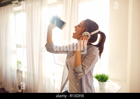 Una ragazza nelle cuffie canta una canzone contro la finestra. Foto Stock