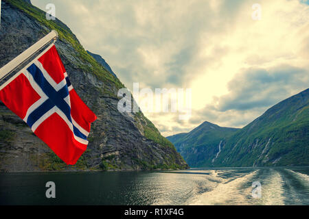 Paesaggio di montagna con cielo nuvoloso. Majestic Geiranger fjord. Vista dalla nave. Bandiera norvegese contro la bellissima natura della Norvegia. Foto Stock