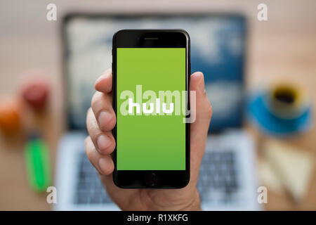 Un uomo guarda al suo iPhone che visualizza il logo di Hulu, mentre se ne sta seduto alla sua scrivania per computer (solo uso editoriale). Foto Stock