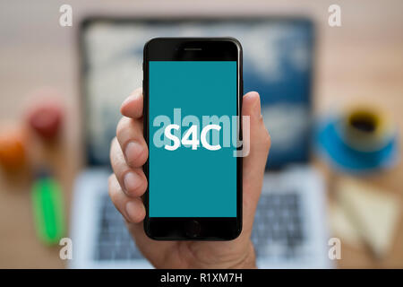 Un uomo guarda al suo iPhone che visualizza la S4C logo, mentre se ne sta seduto alla sua scrivania per computer (solo uso editoriale). Foto Stock