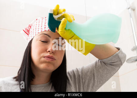 Stanco donna che indossa bandana e tenendo lo spray per la pulizia a mano tocca la fronte come il gemito Foto Stock