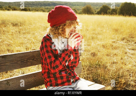 Parte anteriore il ritratto di una giovane donna in rosso plaid shirt con un tappo di lana e sciarpa di prendere una tazza di tè o caffè mentre lei è seduta a prendere il sole in un di legno Foto Stock