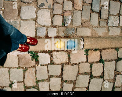 Gambe e piedi di una donna in piedi sul pavimento in pietra. Donna che indossa jeans blu, rosso sneakers e in piedi vicino a giallo e macchie blu Foto Stock