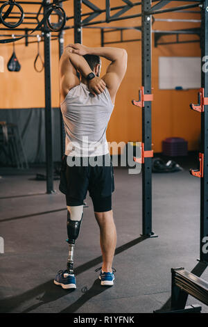 Vista posteriore del giovane sportivo con gamba artificiale stretching vicino alla scaletta di ginnastica in palestra Foto Stock