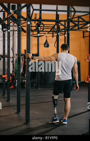 Vista posteriore del giovane sportivo con gamba artificiale in piedi vicino alla scaletta di ginnastica in palestra Foto Stock