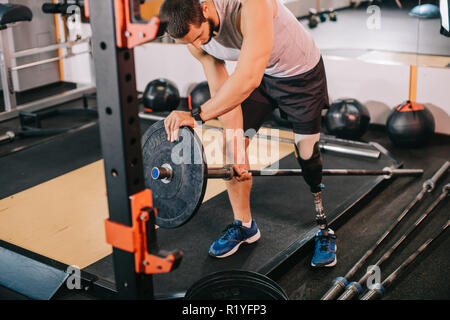 Bel giovane sportivo con gamba artificiale mettendo sulla piastra di peso sulla barra in palestra prima di allenamento Foto Stock