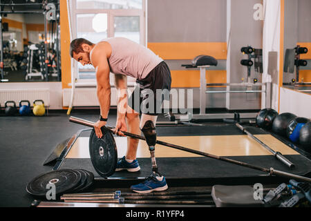 Bel giovane sportivo con gamba artificiale mettendo sulla piastra di peso sulla barra in palestra Foto Stock