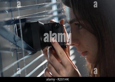 Giovane donna Paparazzi per scattare una foto con sospetto dal intorno a persiane mentre si utilizza una telecamera. Il PILR Concept Foto Stock