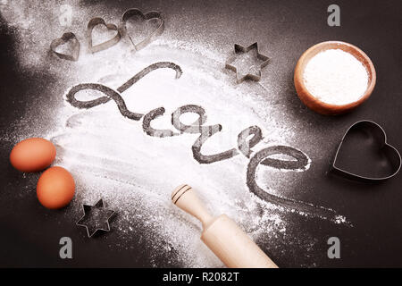 Cuore - conformato cookie cutter, la farina e la parola amore, fatta con esso, e altre cose per i biscotti di cottura, giacente sulla tavola nera. Foto Stock