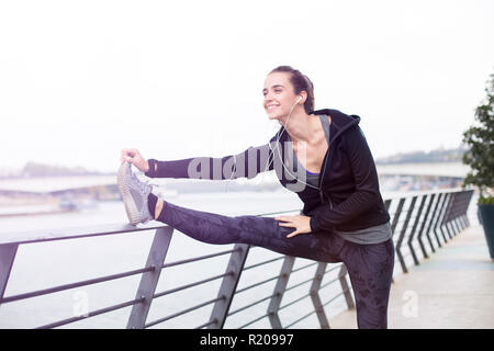 Piuttosto giovane donna con gli auricolari stiramento durante lo sport formazione in ambiente urbano Foto Stock