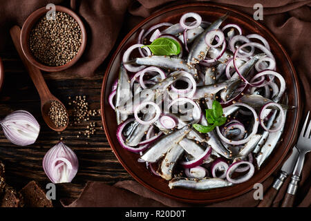 Salate spratti marinato con anelli di cipolle su un piatto di terracotta. Semi di coriandolo, panno marrone, forchetta e coltello su un vecchio rustico tavolo in legno, vista Foto Stock