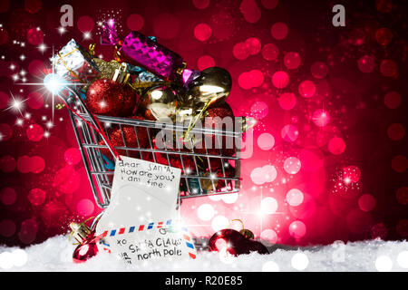 Shopping di Natale concetto, un carrello pieno di regali e una lettera a Babbo natale decorazione, sulla neve, contro una fantastica rosso sfondo bokelicious Foto Stock
