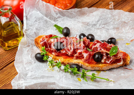 Delizioso pane tostato con pomodoro naturale, olio extra vergine di oliva, il prosciutto iberico, olive nere e le foglie di basilico. Su sfondo di legno. Foto Stock